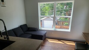 Un espacio habitable compacto con zona de cocina, un sofá de color oscuro, ventana con vista a un patio trasero y suelo de madera.
