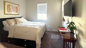 A bedroom with a bed, a desk, a chair, a TV, a window with blinds, and a plant.