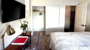 A hotel room with a bed, a desk with a lamp, a TV mounted on the wall, and a closet with a coat hanging.