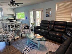 A living room with a black leather sofa, patterned armchairs, a glass coffee table, a dining area with a table and chairs, and a kitchen in the background.