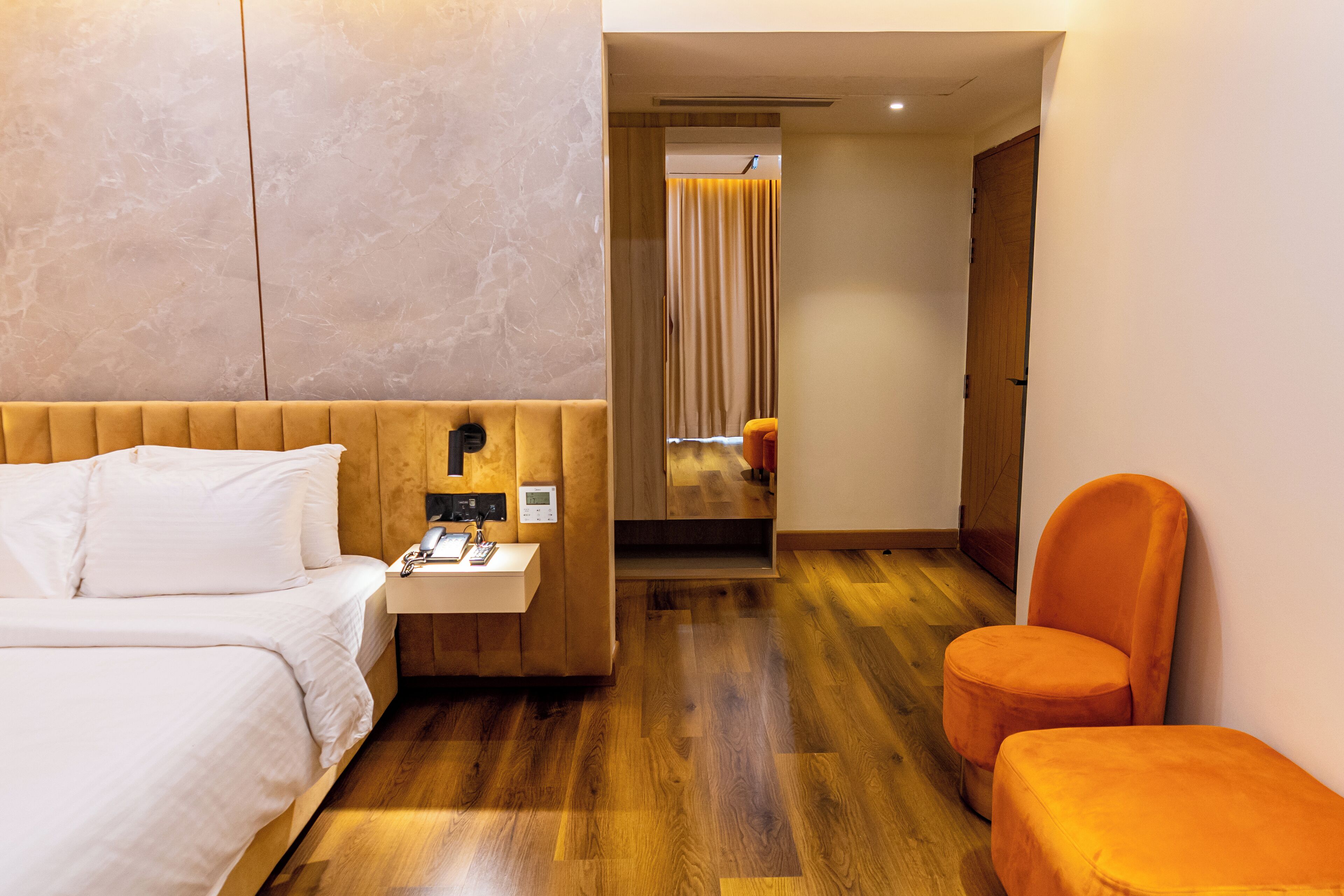 Une chambre d’hôtel avec un plancher en bois, un lit avec du linge de lit blanc, une petite table de chevet avec un téléphone et une lampe, et un fauteuil orange.