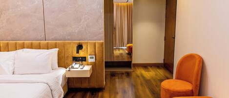 Une chambre d’hôtel avec un plancher en bois, un lit avec du linge de lit blanc, une petite table de chevet avec un téléphone et une lampe, et un fauteuil orange.