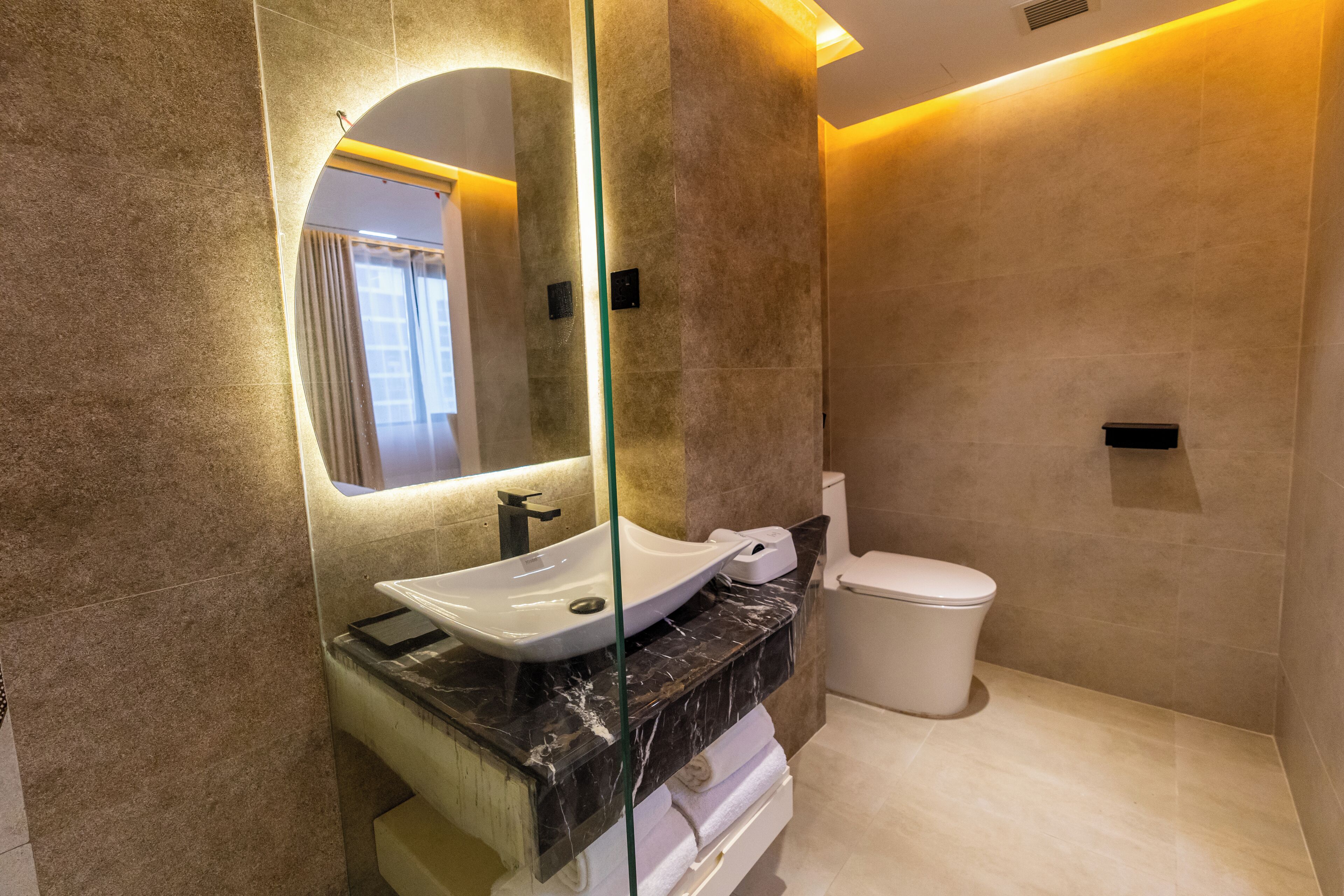 A modern bathroom with a glass-enclosed sink, a marble countertop, a white toilet, and a large mirror.