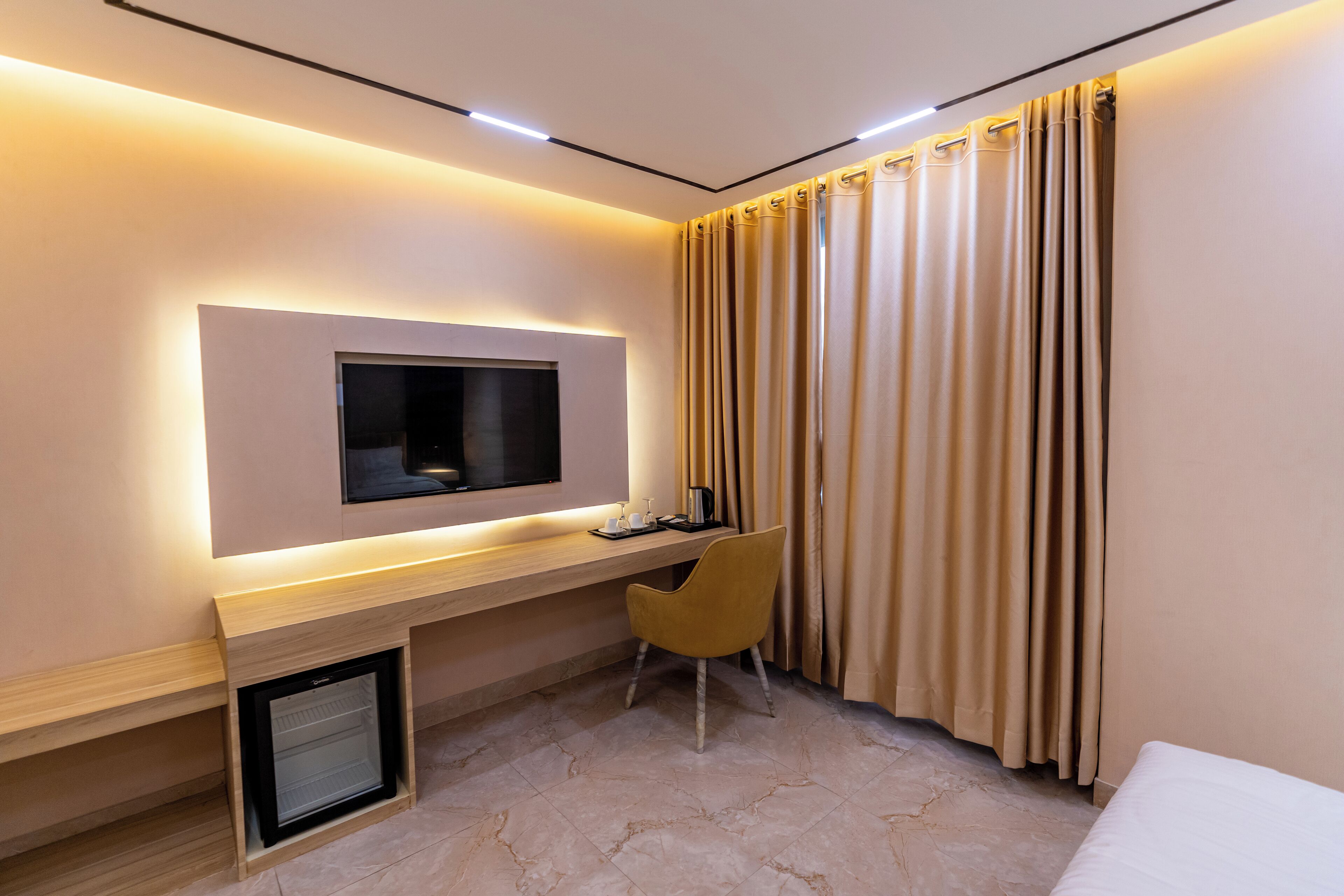 Una habitación de hotel moderna con televisión de pantalla plana en la pared, un escritorio pequeño con una silla, un refrigerador y una cama.