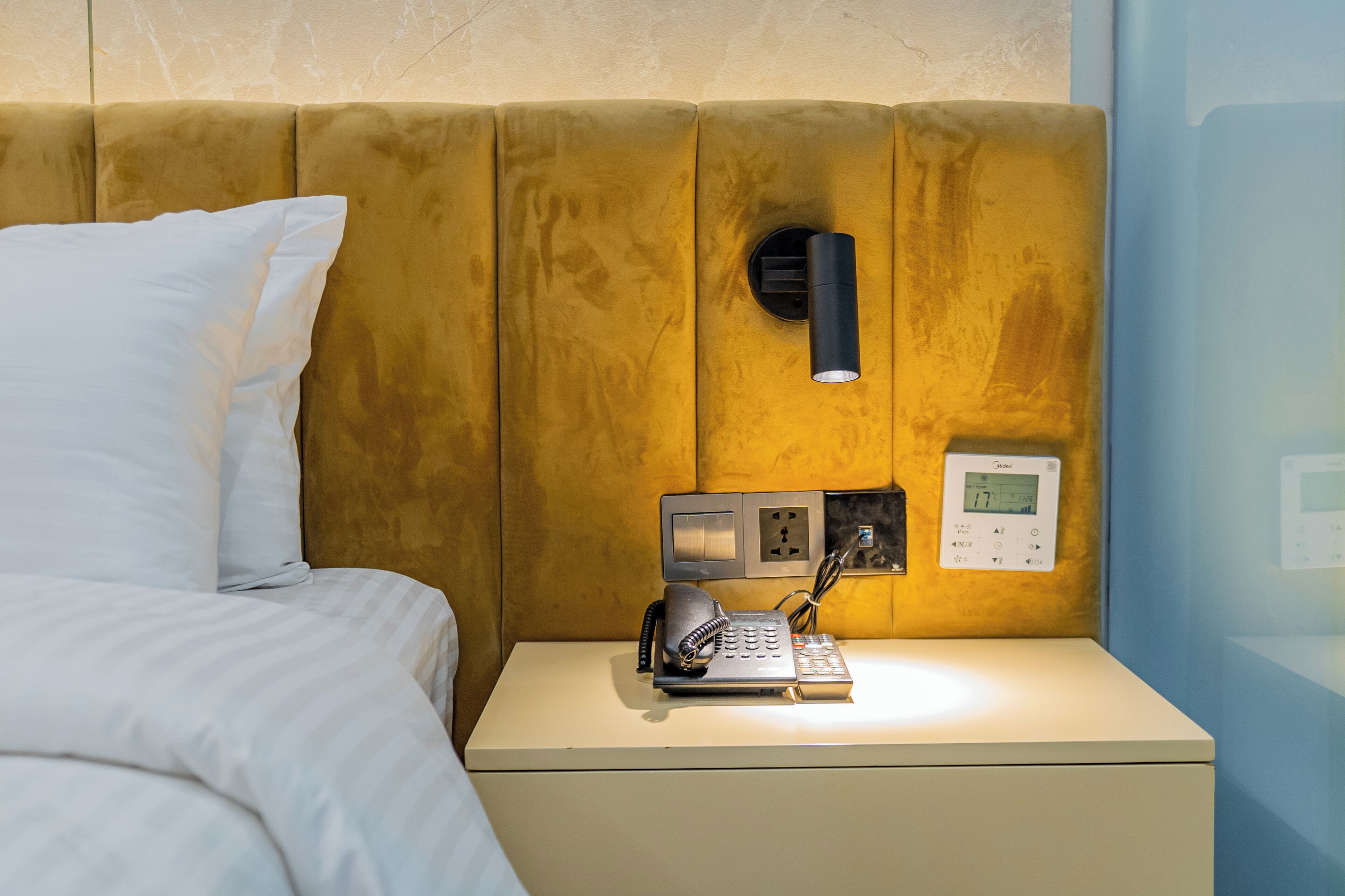 Una habitación de hotel con una cama, una mesita de noche con teléfono, un enchufe de pared y un termostato.