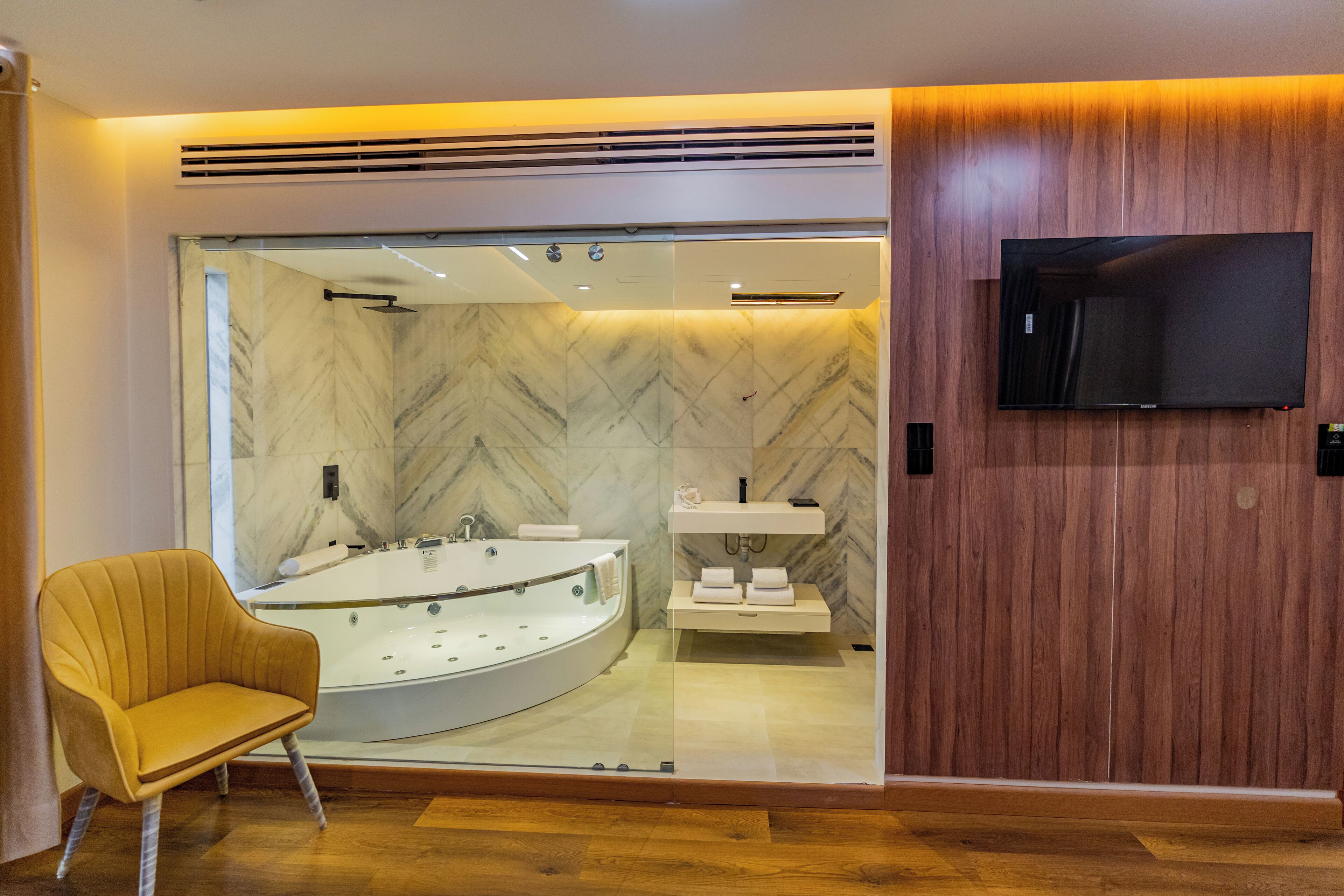 A modern bathroom with a marble wall, a large bathtub, a wooden vanity, and a flat-screen TV mounted on the wall.