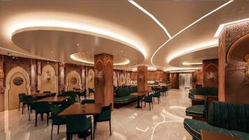 A spacious hotel lobby with a modern design, featuring a central seating area with tables and chairs, and a decorative wall with arches.
