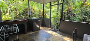 A balcony with a view of dense foliage, a black trash bin, and a metal rack.