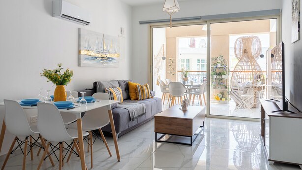 A modern living room with a sofa, dining table, chairs, and a coffee table. There is a large window with a view of the city. - Infinity 1-Bedroom Apartment in Larnaca (Larnaca)