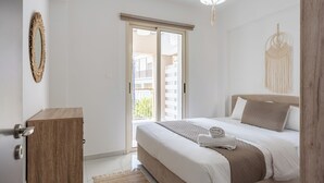 A bedroom with a bed, a wooden dresser, a macrame wall hanging, and a view of the outside balcony. - Infinity 1-Bedroom Apartment in Larnaca (Larnaca)