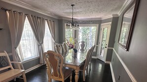 Ein Esszimmer mit einem großen Tisch, umgeben von Stühlen, einer Kandelaber und großen, mit Vorhängen drapierten Fenstern.