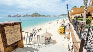 Una playa con una escalera que baja hacia la orilla, un cartel informativo y vistas al mar y colinas distantes.