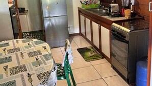 Cocina compacta con refrigerador, estufa y lavamanos. Hay una mesa de comedor con mantel estampado y una silla.