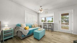 A living room with a blue sofa, a teal ottoman, a ceiling fan, and a window with a view of greenery. - 18+ can book! Panama City Beach, Holiday Beach, The ole MTV house Neighborhood (Panama City Beach)