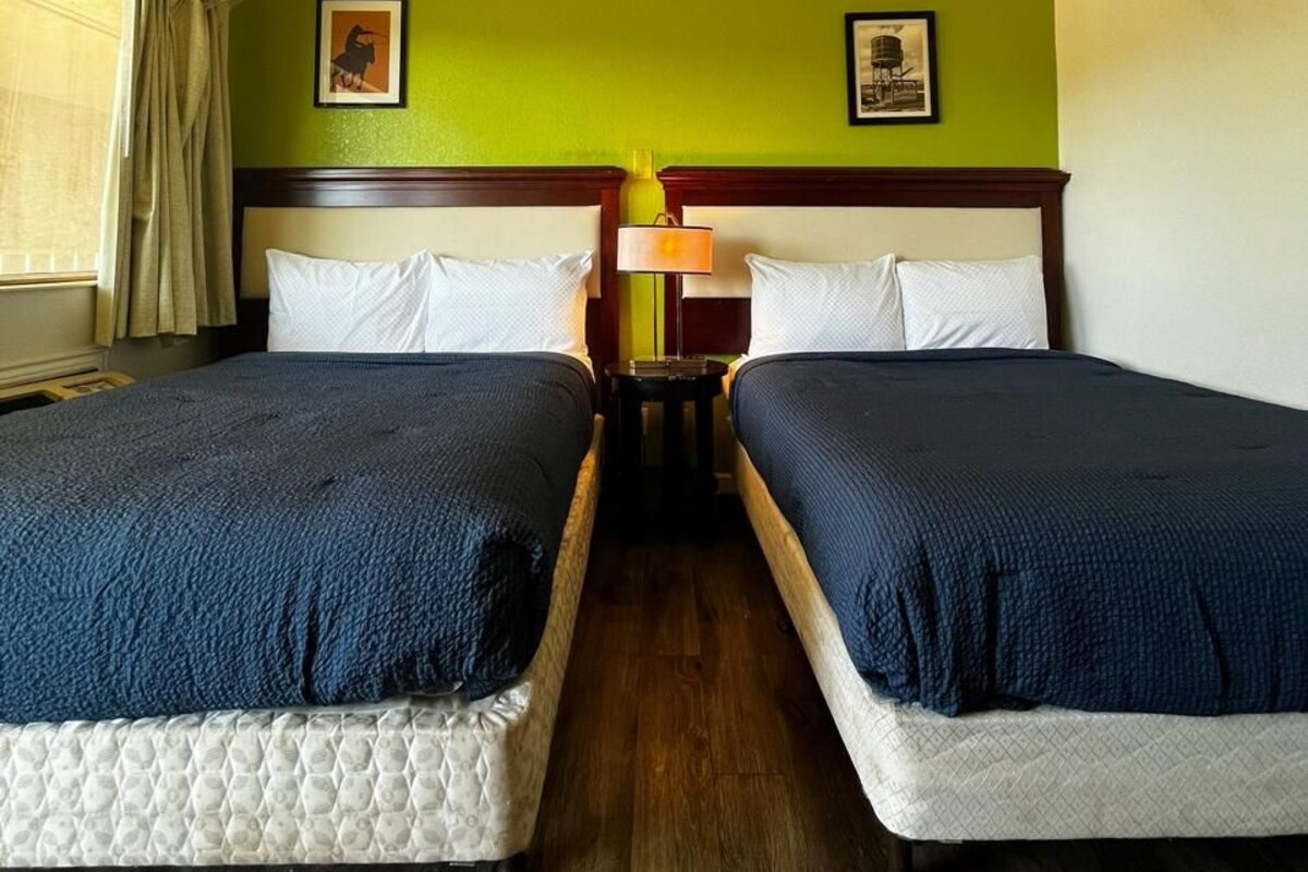 A hotel room with two beds, a wooden headboard, a nightstand with a lamp, and a ceiling light.