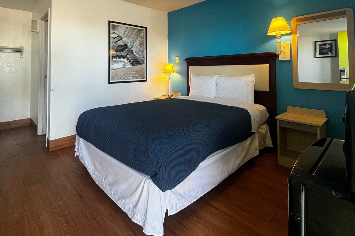 A hotel room with a bed, a desk, a chair, a lamp, a picture on the wall, and a ceiling light.