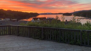 Puinen terassi, jossa on kaide, ja josta avautuu näkymä järvelle, jonka rannalla on taloja ja veneitä, auringonlaskun värittämällä taivaalla.