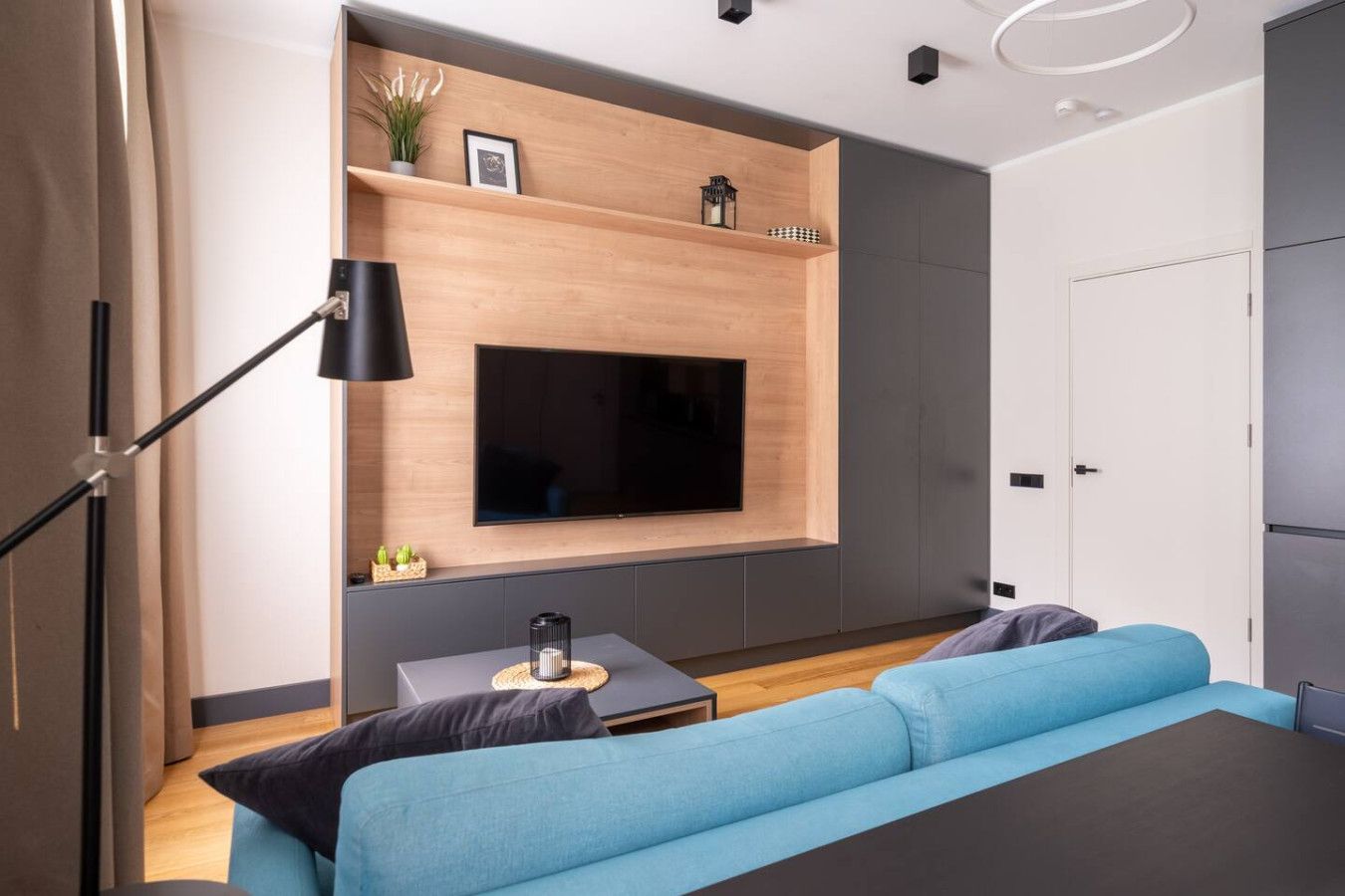 A modern bedroom with a large TV mounted on a wooden wall, a blue sofa, a small side table, and a floor lamp.