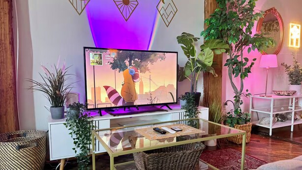 A modern living room with a flat-screen TV on a white stand, surrounded by plants, a wicker coffee table, and a red patterned rug. - Downtown: River’s Edge Industrial Loft (Detroit)