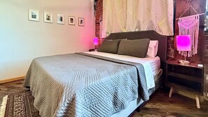 A bedroom with a bed, a nightstand, a wall-mounted macrame piece, and framed pictures on the wall. - Downtown: River’s Edge Industrial Loft (Detroit)