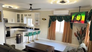 A modern living room with a sofa, a coffee table, and a TV. There is a kitchen area with white cabinets and a black countertop. - Charming 3-bedroom villa in Ocho Rios with a view of the Caribbean Sea (Ocho Rios)