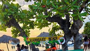 A beach scene with a sign reading "Mahogany Beach Bar & Grill" under a tree. There are colorful benches and a thatched umbrella. - Charming 3-bedroom villa in Ocho Rios with a view of the Caribbean Sea (Ocho Rios)