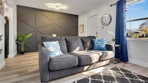 Una sala de estar moderna con un sofá gris, almohadones azules estampados, una alfombra blanco y negro, un reloj de pared y una ventana con cortinas azules.