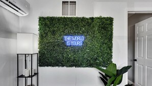 Una habitación con una pared verde cubierta de follaje, un letrero de neón que dice "THE WORLD IS YOURS", una puerta blanca y una planta a la derecha.