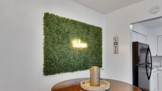 Zona pranzo con un tavolo rotondo in legno, una parete verde decorata con piante, un neon e una cucina sullo sfondo.