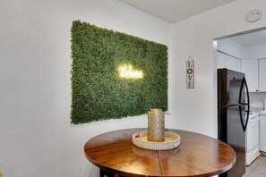 Un espace repas avec une table ronde en bois, un mur végétal vert fixé au mur, un panneau lumineux au néon et une cuisine en arrière-plan.