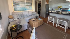 A modern living room with a grey sofa, a coffee table, and a kitchen area with a refrigerator and sink.
