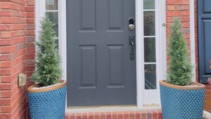 Una puerta delantera gris oscura con un aldabón dorado, flanqueada por dos macetas azules con pequeños árboles, y un felpudo con un borde decorativo.