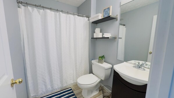 Un baño con inodoro y lavamanos blancos, cortina de ducha, toallero y un estante pequeño con artículos de aseo.