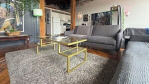 A modern living room with a grey sofa, a glass coffee table, and a kitchen in the background. - Downtown: Cinema Loft w/ Projector (Detroit)