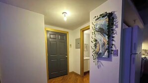 A hallway with wooden flooring, a dark door, a refrigerator, and a wall-mounted artwork. - Downtown: Cinema Loft w/ Projector (Detroit)