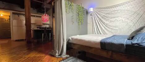 A bedroom with a bed, a hanging plant, a patterned wall, and a kitchen in the background.