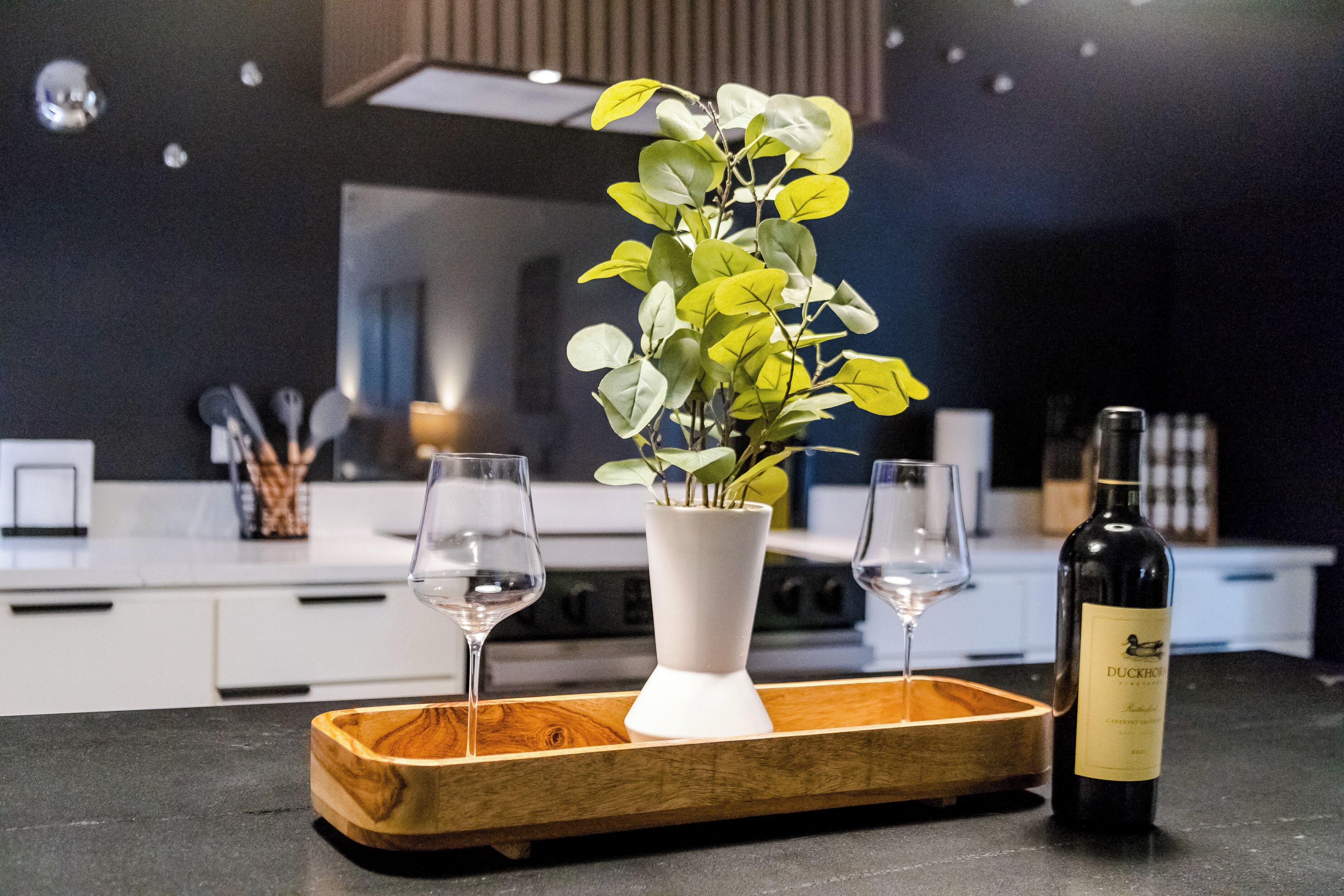 A modern kitchen with a white vase containing green leaves, two wine glasses on a wooden tray, and a bottle of Duckhorn wine on the counter.
