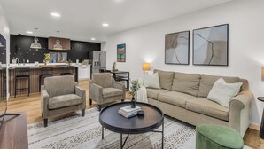 En moderne stue med sofa, lænestole og sofabord. Et køkken med spiseplads ses i baggrunden.