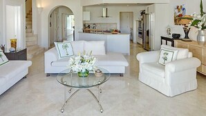 A modern living room with a glass coffee table, white sofas, and a kitchen in the background. - Schooner Bay 306 - Penthouse  | Beach Front - Located in  Exquisite St. Peter with House Cleaning Included (Speightstown)