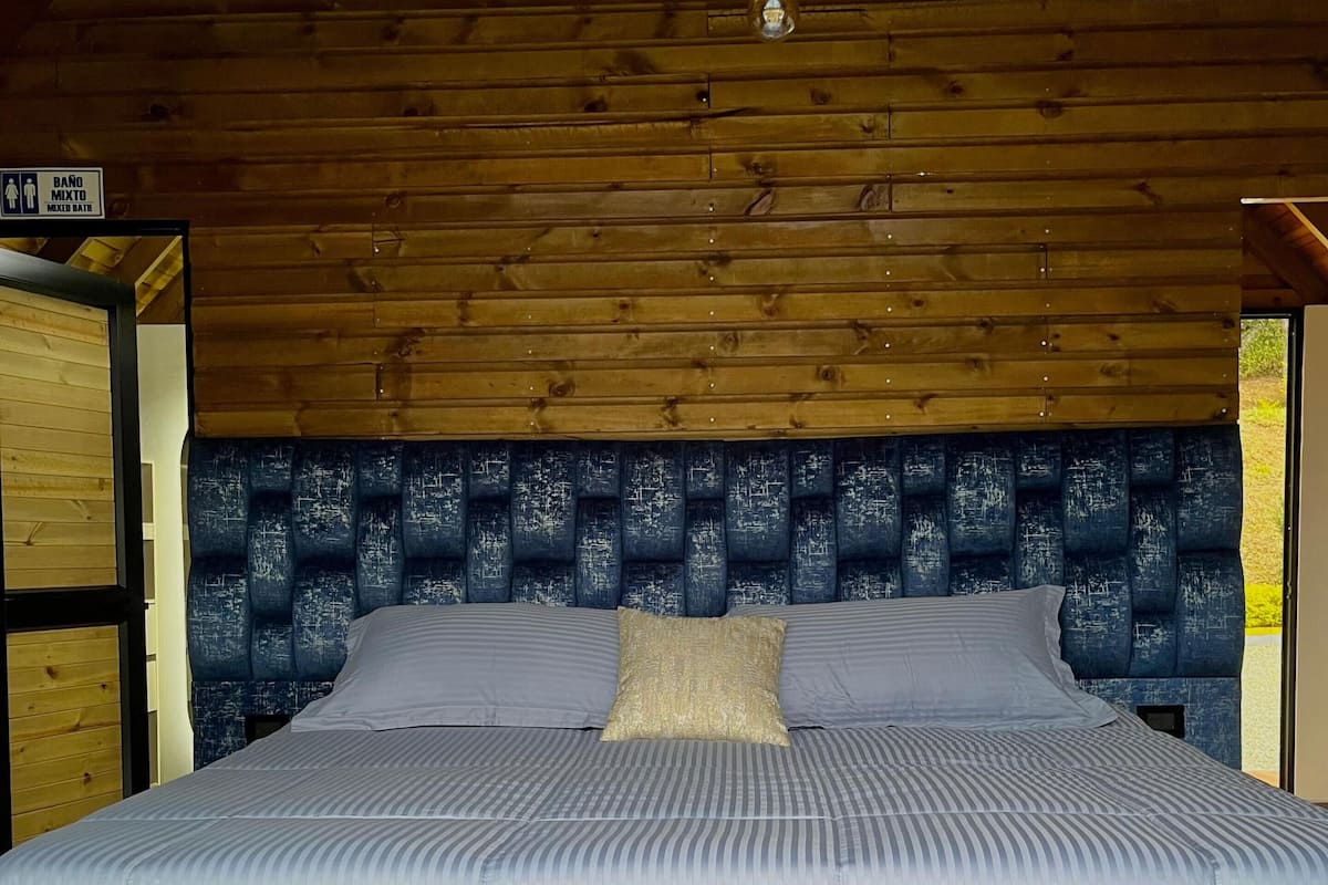 A bedroom with a wooden ceiling, a bed with a blue headboard, and a sign indicating a mixed bath.
