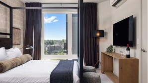 A modern hotel room with a large bed, a desk with a TV, a bottle of wine, and a view of the city. - Luxury Apartments by Hyatus White Plains (White Plains)