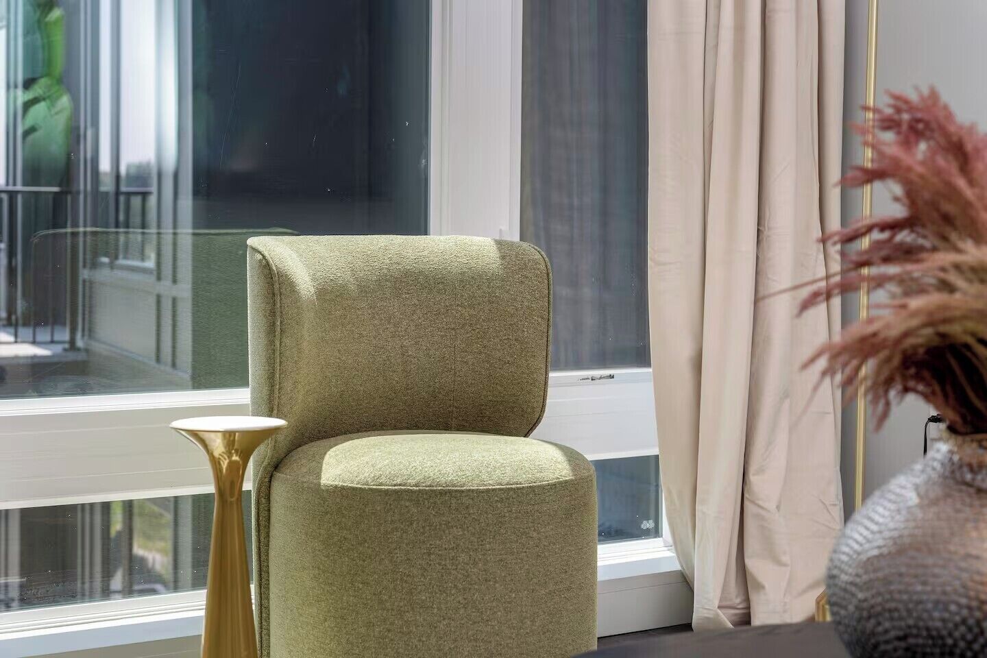 A room with a green upholstered chair, a small round table, and a vase with dried plants.