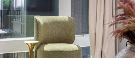 A room with a green upholstered chair, a small round table, and a vase with dried plants.
