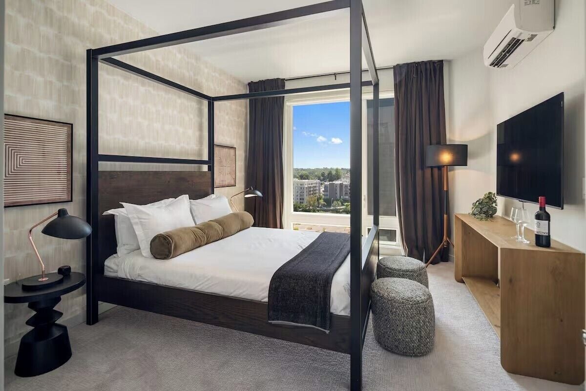 Une chambre d’hôtel moderne dotée d’un lit à baldaquin, d’un bureau avec une télévision, d’une grande fenêtre avec des rideaux et d’un climatiseur fixé au mur.