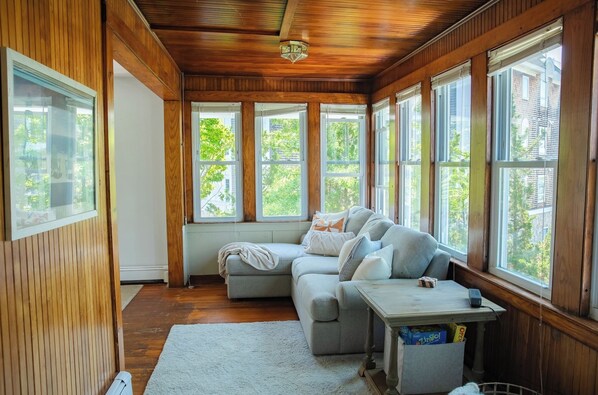 Una habitación acogedora con techo de madera, ventanas amplias, un sofá seccional gris y una mesita pequeña.