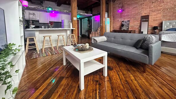 A modern living room with a grey sofa, wooden flooring, and a kitchen area with bar stools. - Downtown: Industrial Brick Chill Loft (Detroit)