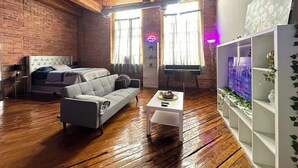 A room with a brick wall, wooden ceiling, a bed, a grey sofa, a small white table, and a white shelving unit with decorative items. - Downtown: Industrial Brick Chill Loft (Detroit)