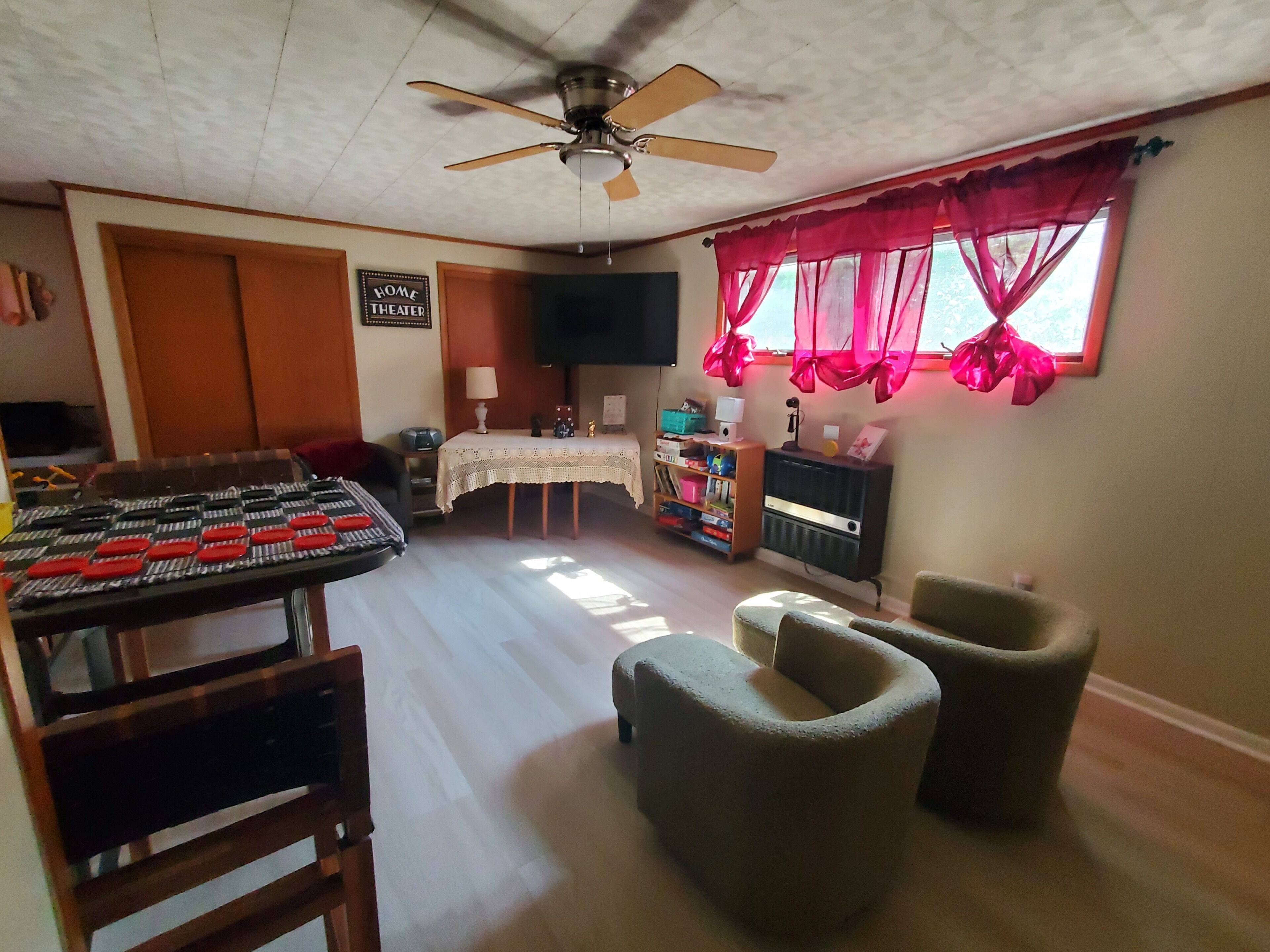 A living room with a ceiling fan, a table with a lace tablecloth, a television, and a window with pink curtains.