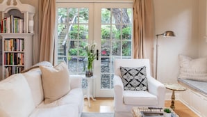A cozy living room with a white sofa, a patterned armchair, a bookshelf, and a view of a garden through a glass door. - Charming 1-bedroom cottage in gorgeous Monterey (Monterey)