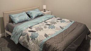 Ein ordentlich bezogenes Bett mit gemusterten Betttextilien, ein Nachttisch mit Lampe und ein Fenster mit Jalousien.
